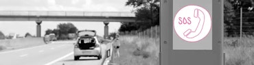 Notrufsäule an einer Autobahn – im Hintergrund ein liegengebliebenes Auto und Person in Warnweste.