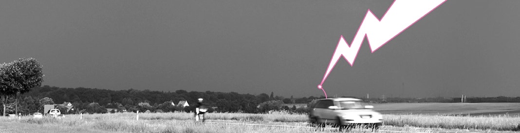 Blitzeinschlag bei Gewitter: So sicher ist Ihr Auto Ein Auto fährt im Regen unter dunklen Gewitterwolken – sicher vor Blitzeinschlag durch die metallische Karosserie als Faradayscher Käfig.